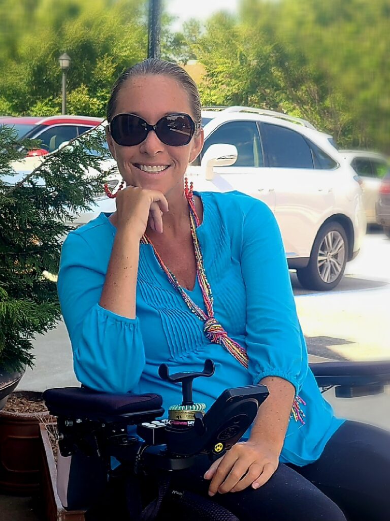 picture of Ali Ingersoll and wheelchair from waste up with hand on chin smiling and sunglasses and bright blue top