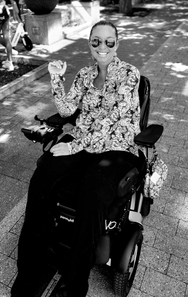 picture of Ali Ingersoll in wheelchair and black-and-white with sunglasses and bright top on with black pants