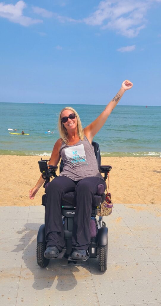 Ali and wheelchair with beach in the background arms waving in the air