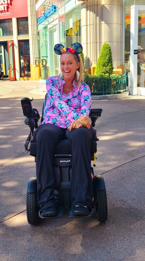 image of Ali and wheelchair at Disneyland with Disney years on in bright neon top and black pants