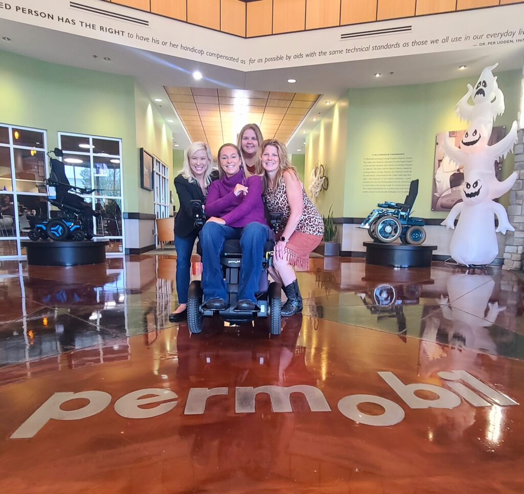 Ali and permobil front hallway of factory with four other women smiling behind Ali and wheelchair
