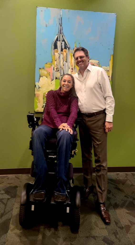 image of Ali and wheelchair in blue jeans and purple sweater standing next to Chuck Witkowski, president of permobil Americas with dark green wall background with painting and back smiling