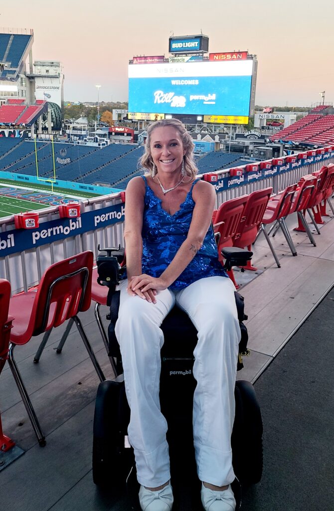 Ali in wheelchair with spaghetti strap blue sequence top with white pants with Nissan Stadium in Nashville Tennessee in the background