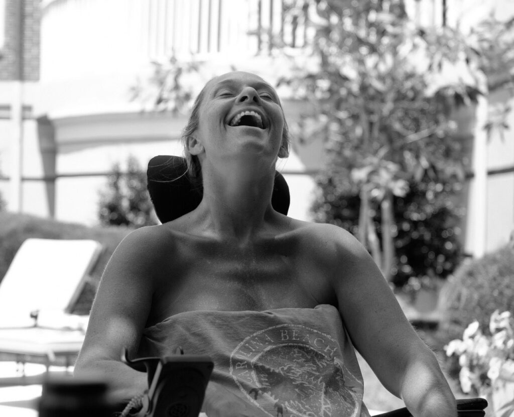 image of Ali from waste up in wheelchair in black-and-white with head tilted back laughing