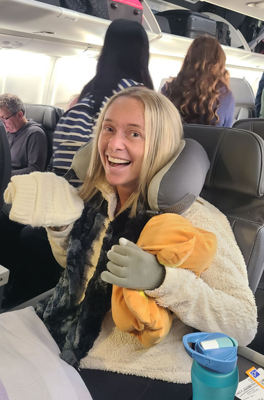 Ali in white fluffy fleece with pillows with neck wrap in airline seat with blonde hair making funny face