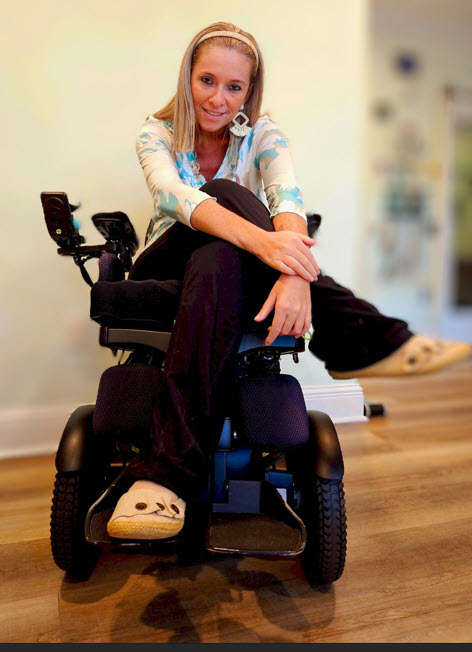Ali and wheelchair leaning forward with black pants and blue-and-white shirt smiling with long blonde hair and big beige Earrings with headband