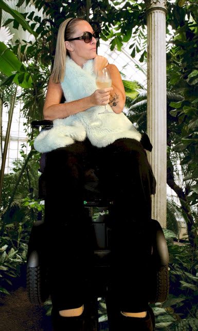 Ali and wheelchair in black tank top and black pants with blue scarf wrapped around neck with glass of wine.  Blonde hair, sunglasses with tropical background